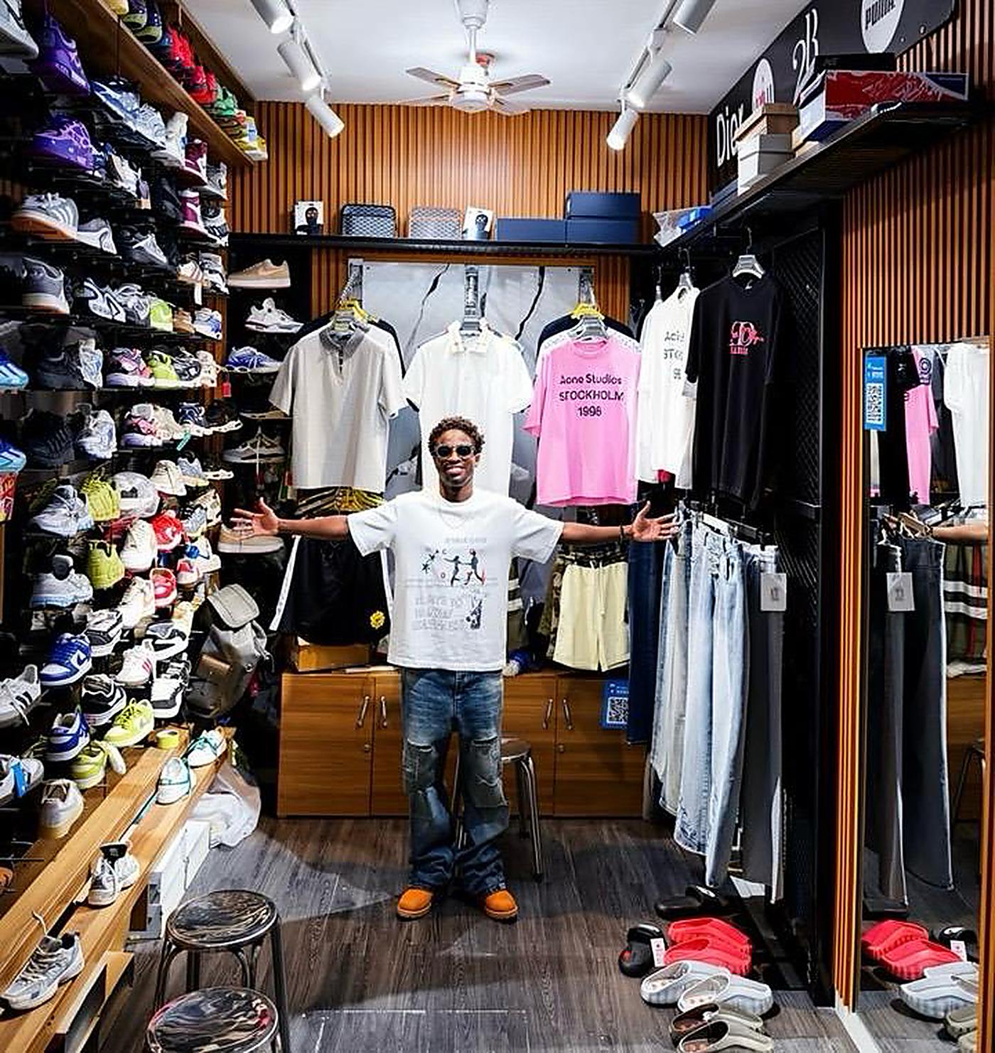 Un vendeur dans sa boutique à Dakar, bras ouverts entre un mur de sneakers et ses portants de vêtements.
